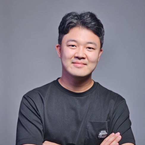 Professional portrait of a person with short dark hair wearing a black t-shirt with a small pocket design. They have their arms crossed and are slightly smiling at the camera against a gray background. The person is wearing a dark watch on their wrist. 
