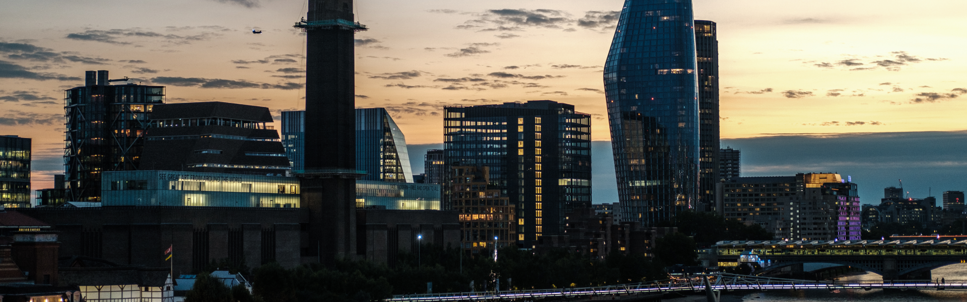 City skyline at sunset with modern glass buildings, a tall brick smokestack, and a bridge with illuminated lights crossing a river in the foreground.