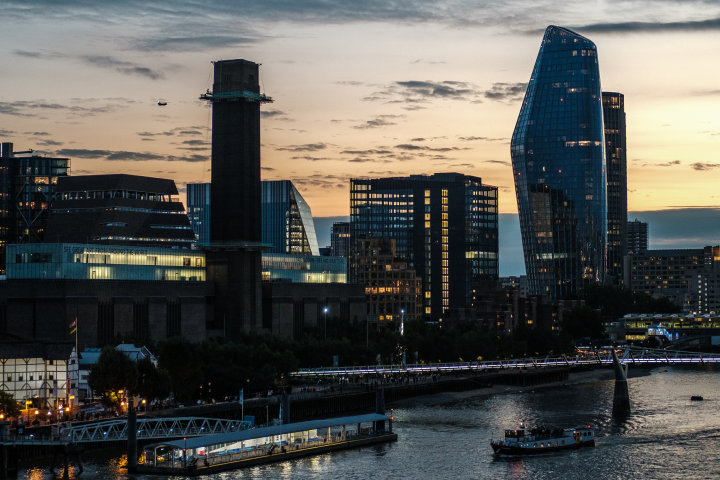City skyline at sunset with modern glass buildings, a tall brick smokestack, and a bridge with illuminated lights crossing a river in the foreground.